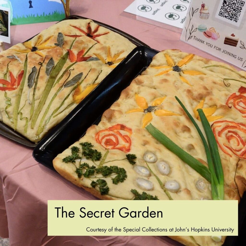 Image description: Two trays of focaccia bread with vegetables baked into the top to look like flowers, leaves, and other plants. In the bottom right, a green rectangle overlays the picture with black text, reading "The Secret Garden, Courtesy of the Special Collections at John's Hopkins University."